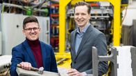 Kaksi pikkutakkeihin pukeutunutta miestä hymyilee betonitehtaalla, betonipalkkien välissä.