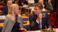 A woman with blonde hair speaking to a man with brown hair who is holding a cup, while both are seated at a table.