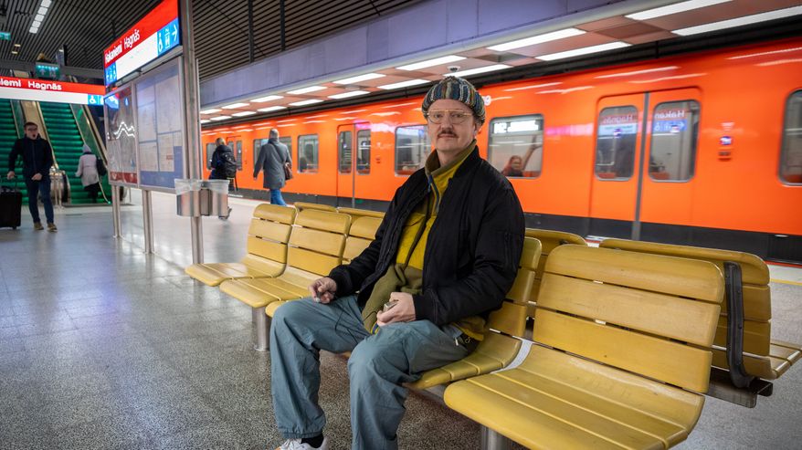 Mies istuu hakaniemen metroaseman puisilla penkeillä ja ympärillä vilisee ihmisiä ja metro taustalla.