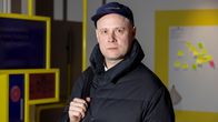 Huoneessa seisoo nuori mies, jolla on sininen takki ja sininen lippalakki.