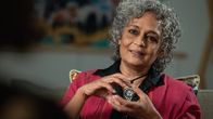 Arundhati Roy elehtii käsillään puhuessaan. Hän on pukeutunut punaiseen takkiin ja mustaan kauluspaitaan. Kaulassa on riipus.