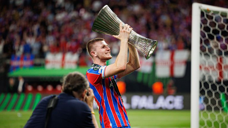 En fotbollsspelare i rödblå randig tröja lyfter en stor pokal, en fotograf tar bild framför honom.