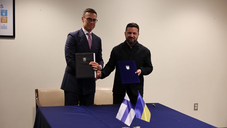 Kaksi miestä seisoo pöydän ääressä pitäen kansioita ja kättelee, pöydällä Suomen ja Ukrainan liput.