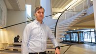 Nuorehko mies seisoo rakennuksen aulassa kierreportaiden juuressa ja nojaa toisella kädellään kaiteeseen.