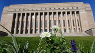 Eduskuntatalo Helsingissä, etualalla ruohoa ja kukkia, taustalla sininen taivas.