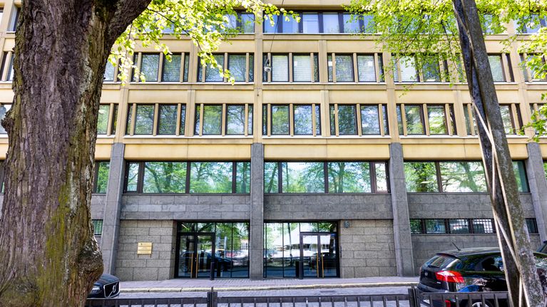 Facade of a beige and grey office building framed by trees with green leaves, with a black car parked in front.