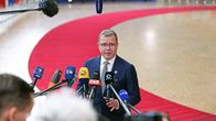 En man i kostym står framför flera mikrofoner och bandspelare på stativ; bakom honom syns en röd matta och flaggor.