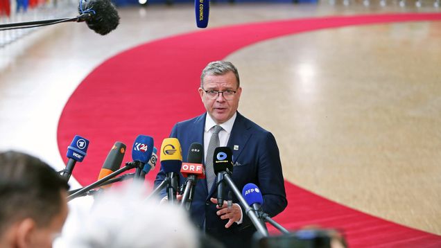 Mies seisoo mikrofonien edessä antamassa lausuntoa, taustalla punainen matto ja useita maiden lippuja.