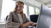 Nainen istuu toimistossa puhelinkuullokkeet päässä ja kirjoittaa kannettavalla tietokoneella.
