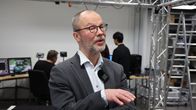 Silmälasipäinen mies harmaa puvuntakki päällään seisoo huoneessa. Taustalla ihmiset työskentelevät tietokoneella. 