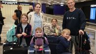 A father, mother and four young kids pose for a photo amid a large number of suitcases and bags at an airport arrivals hall.