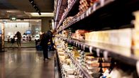 Person wearing black stooping over a grocery store refrigerated goods shelf.