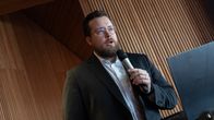 En person i kostym håller i en mikrofon och står bredvid en bärbar dator framför en vägg av träpanel.