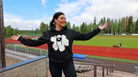 Nainen mustassa Marimekon hupparissa levittelee iloisena käsiään. Taustalla urheilukenttä.