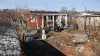 Pieni punainen mökki, jonka edustalla kaksi henkilöä seisoo puutarhassa.