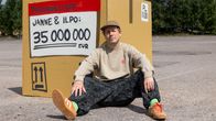 Mies seisoo asfalttialueella ison laatikon vieressä, jossa lukee ”PAKKAUSLISTA Janne & Ilpo: 35 000 000 EUR”. Taustalla puita.