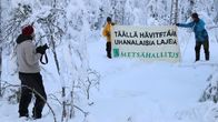Kaksi henkilöä levittää banderollia lumisessa metsässä, valokuvaaja seisoo vieressä.