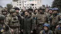 Ukrainan presidentti Volodymyr Zelenskyi seisoo sotilasjoukon edessä. Taustalla näkyy tuhoutunut kerrostalo.