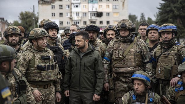 Ukrainan presidentti Volodymyr Zelenskyi seisoo sotilasjoukon edessä. Taustalla näkyy tuhoutunut kerrostalo.