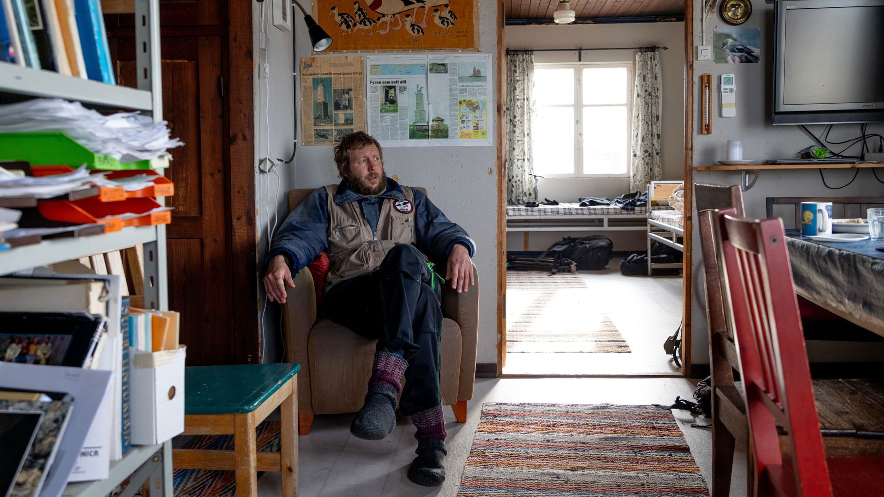 Antti Miettunen-Nordström istuu nojatuolissa vanhan tuvan huoneen nurkassa. Vasemmalla kirjahylly, lattialla räsymatto ja oikealla ruokapöydän tuoleja. 
