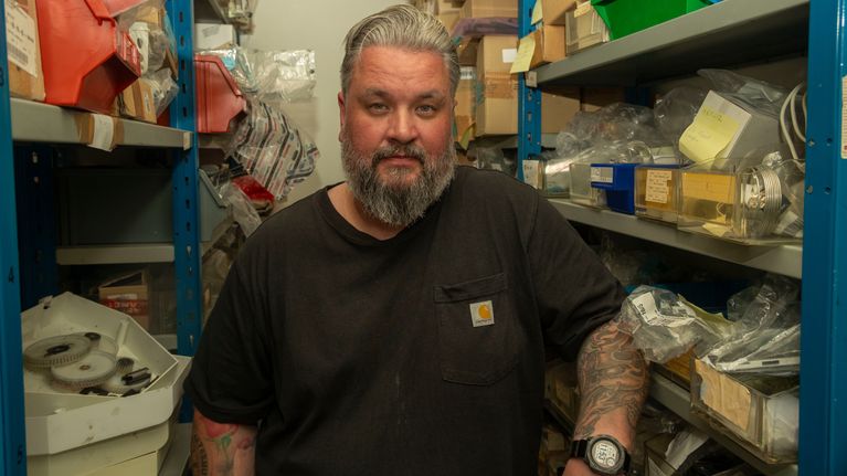 Mies seisoo varastossa erilaisten tarvikelaatikoiden ympäröimänä. Hänellä on päällään musta Carhartt-paita ja ranteessaan kello. Seinustalla näkyy teknisiä osia ja paketteja hyllyissä.