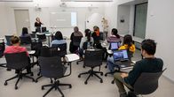 About 10 people, mostly women, two wearing headscarves, seen from behind as they sit in a classroom with a female teacher standing in front.