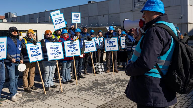 Joukko ihmisiä seisoo kadulla mielenosoituskylttien kanssa ja yksi henkilö heidän edessään pitelee megafonia.