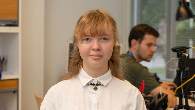 Vaaleahiuksinen nainen katsoo kameraan. Taustalla pöydän ääressä istuu toinen henkilö tietokoneella.
