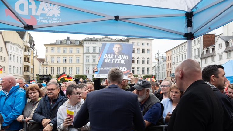 Ihmismassa vanhojen rakennusten reunustamalla torilla, kuvattu vaaliteltan alta selin kameraan olevien miesten takaa.