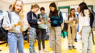 En grupp flickor står i ett rum och tittar på de mobiltelefoner de har i händerna. De verkar vara i en skola eftersom de har ryggsäckar hängande på axeln.