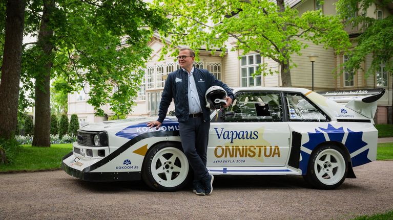 Petteri Orpo poseeraa ralliauton edessä kypärä kainalossa. Autossa lukee "vapaus onnistua". Taustalla vanha hieno puurakennus, Kesäranta.