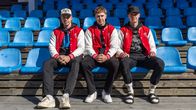 Kolme miestä samanlaisissa takeissa istuu katsomon sinisissä penkeissä.