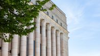 Green tree branches in front of a large off-white neoclassical building with columns and a blue sky. 