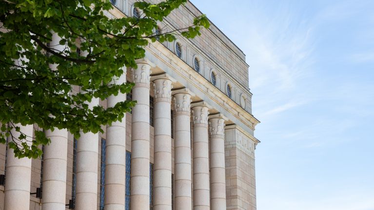 Green tree branches in front of a large off-white neoclassical building with columns and a blue sky. 