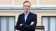 En man står framför en byggnad, klädd i en mörk kostym och blå skjorta, med armarna över kors. I bakgrunden syns fönster med persienner och en träbänk vid den gulmålade fasaden.