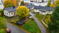 Ilmakuva syksyisestä pihapiiristä, jossa on kaksi vanhaa vaaleaa puutaloa ja puustoa ympärillä, etualalla kaksi autoa.