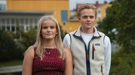 vå unga personer står utomhus framför gula byggnader, omgivna av grönska.