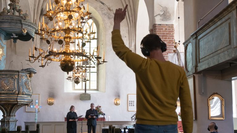 En person med hörlurar står med armen lyft i en kyrka där två präster står framför altaret.