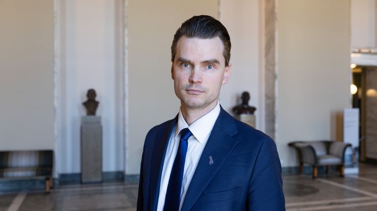 Photo of a man in dark blue suit jacket, white shirt and dark blue necktie.