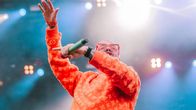 Oranssiin paitaan ja punaisiin aurinkolaseihin pukeutunut J Balvin kädet ilmassa Ruisrockin Niittylavalla. Kuva rajattu vyötäröstä alaspäin.