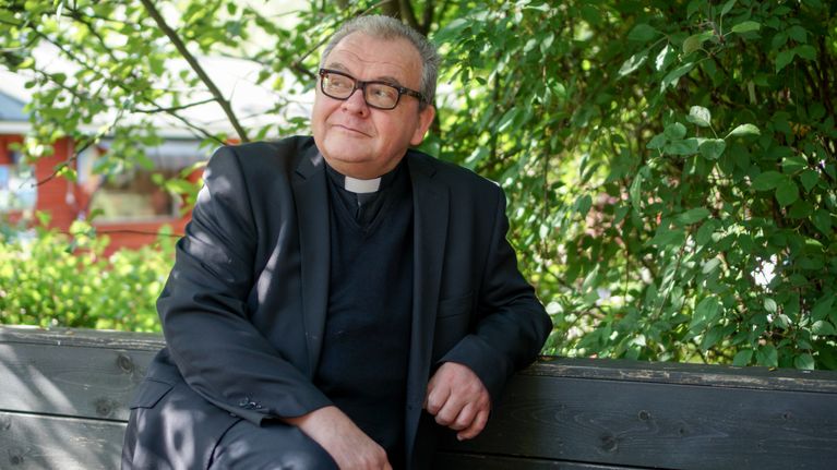En man i prästkappa och prästkrage sitter utomhus på en bänk, omgiven av gröna träd och buskar.