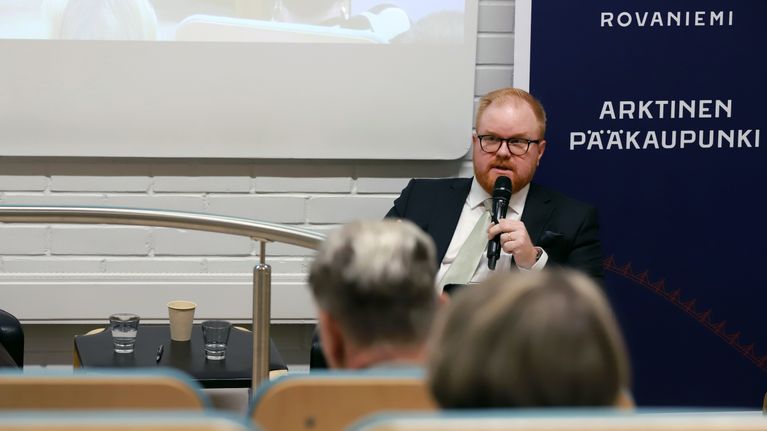 Mies tummassa puvussa istuu puhumassa mikrofoniin.