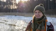 Bilden visar ett vinterlandskap med snötäckt mark och skog i bakgrunden. Kvinna i förgrunden som ler in i kameran.
