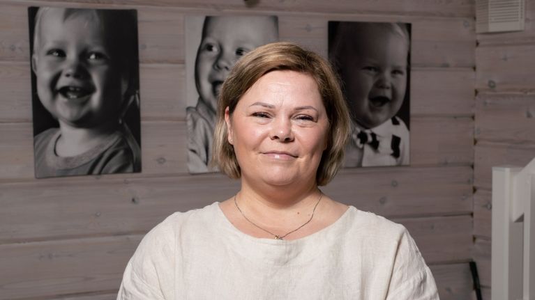 Nainen katsoo kameraan, taustalla valokuvia lapsista.