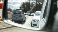 Liikenteen ruuhkaa kuvattuna auton sivupeilin kautta, jossa näkyy etualalla henkilöautoja ja taustalla linja-auto, ja kadun varrella kulkeva kaaritie.