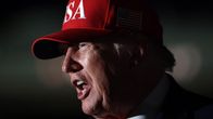 Donald Trump punaisessa lippiksessä, jossa lukee "USA" ja Yhdysvaltain lippu, puhuu tummassa ympäristössä.