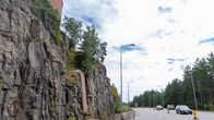 Valtatiellä kulkee useita autoja. Tien vieressä on suuri kallioseinä jossa on putkia. Kallion päällä on vesitorni.