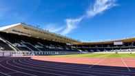 Tyhjä yleisurheilustadion, jonka juoksuratakaaret näkyvät etualalla ja katsomot taustalla. Selkeää taivasta.