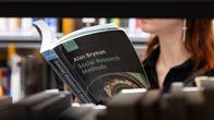 Henkilö valitsemassa kirjaa kirjaston hyllyltä; kädessään Alan Brymanin teos "Social Research Methods".