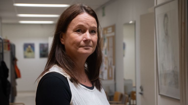 Annelie Åkerman-Anttila har brunt hår och står i en korridor med konstverk och anslagstavlor på väggarna. Hon bär en svart och vit topp.